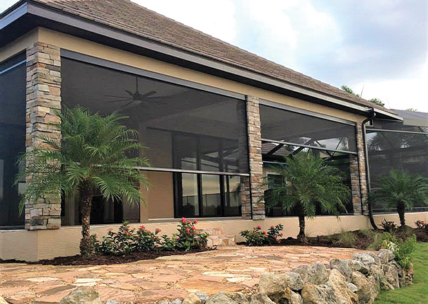 A covered patio exterior with stone columns and openings protected by retractable screens in a dark color.

