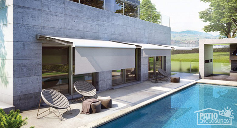 A narrow patio behind a pool with several chairs and two shie retractable awnings with drop valances extended.  