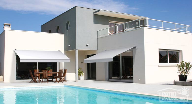 A white and gray contemporary home with an in-ground pool and outdoor patio shaded by two retractable awnings.