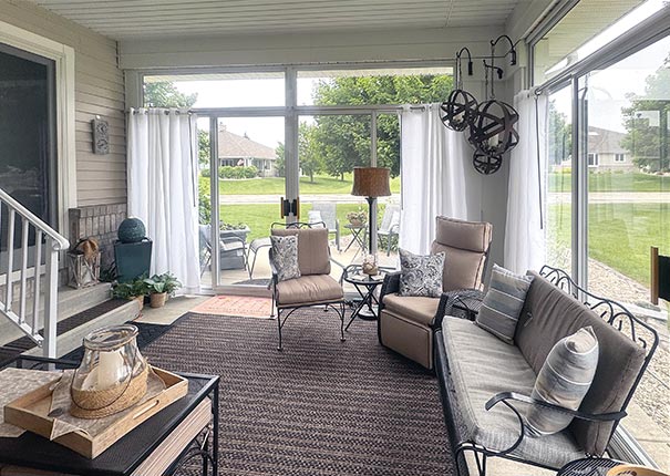 A corner patio enclosure glass sunroom with comfortable furnishings, white sheer drapes, and hanging lamps in the corner.
