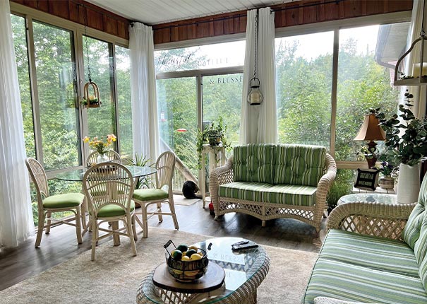 A glass sunroom with sheer drapes, green striped cushions on white wicker furniture, round glass table and chairs.
