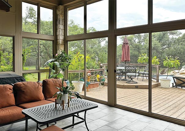 A screened sunroom with tall walls overlooking a deck with table and chairs. Inside an orange sofa, coffee table, and plants.
