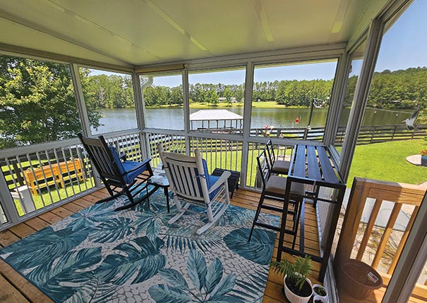 Second-story porch enclosure sunroom overlooking a large lawn and lake. Furnished with rocking chairs, long table and chairs.
