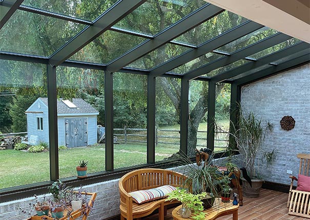 A brown solarium sunroom with brick knee wall, glass ceiling and wood floor. Furnished with wood furniture and plants.
