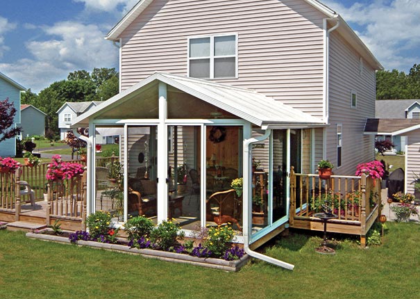 A white gable-roofed glass sunroom kit with attached deck and colorful flowers around on a beige colonial home.

