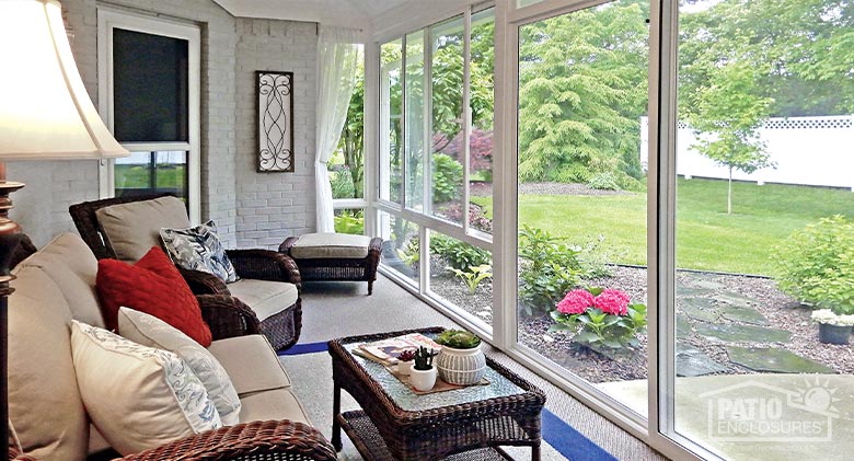 A backyard view from the interior of a glass-enclosed porch or lanai furnished with cushioned wicker furniture and pillows.